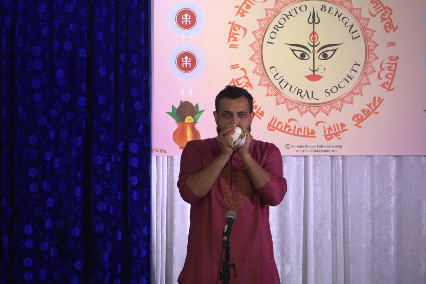Man blowing a conch shell in front of the Toronto Bengali Cultural Society banner